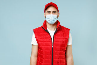 Delivery guy employee man in red cap white t-shirt vest waistcoat uniform sterile face mask gloves work courier service on lockdown coronavirus virus looking camera isolated on pastel blue background