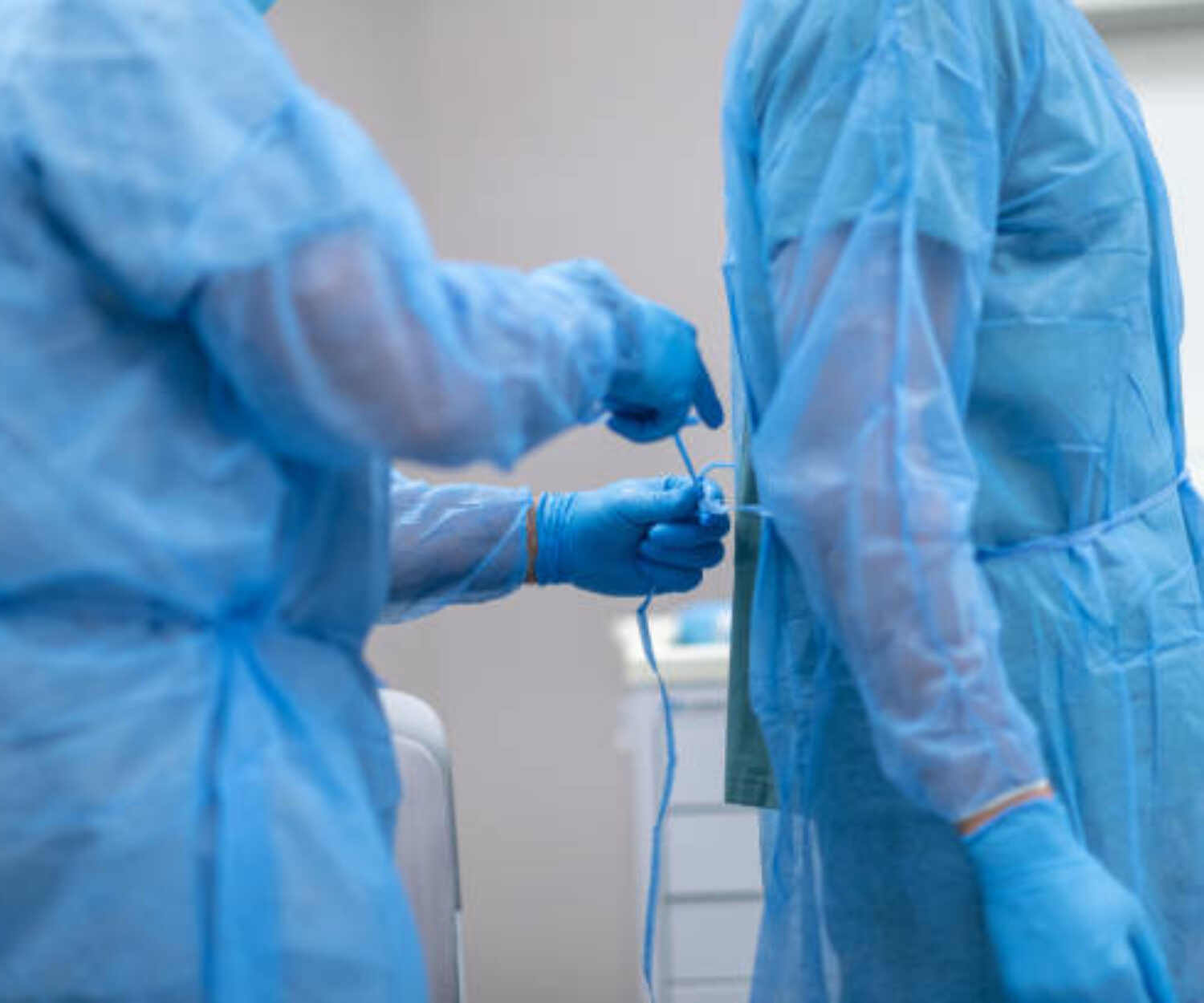 Two surgeons are preparing for surgery. The doctors are standing in an operating room in a hospital. One surgeon is tying closed the back of another surgeon's operating gown.