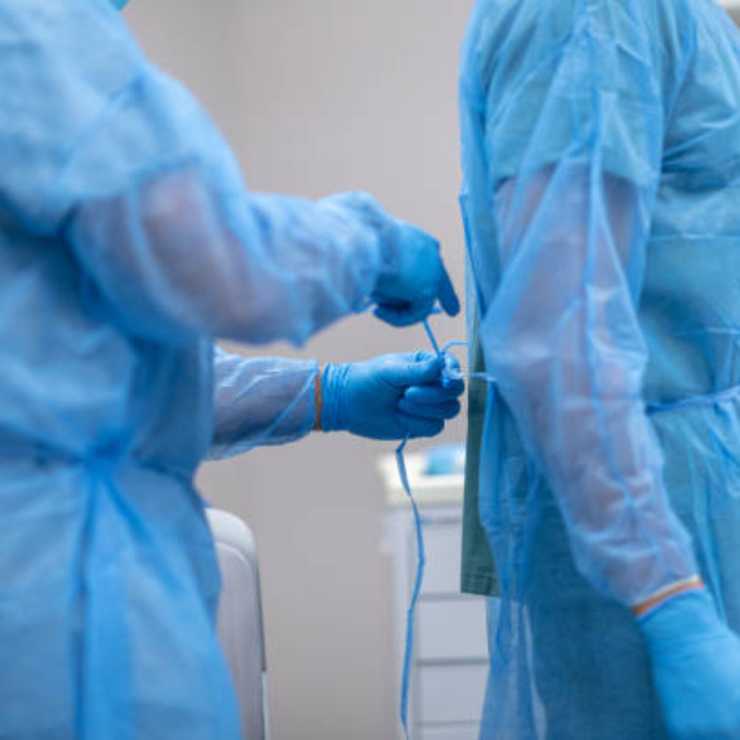 Two surgeons are preparing for surgery. The doctors are standing in an operating room in a hospital. One surgeon is tying closed the back of another surgeon's operating gown.