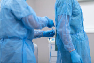 Two surgeons are preparing for surgery. The doctors are standing in an operating room in a hospital. One surgeon is tying closed the back of another surgeon's operating gown.